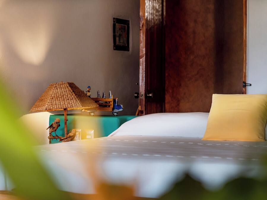 A neatly made bed with a white comforter and a single yellow pillow, a small wooden nightstand with a lamp and a figurine, and a wooden door in the background.