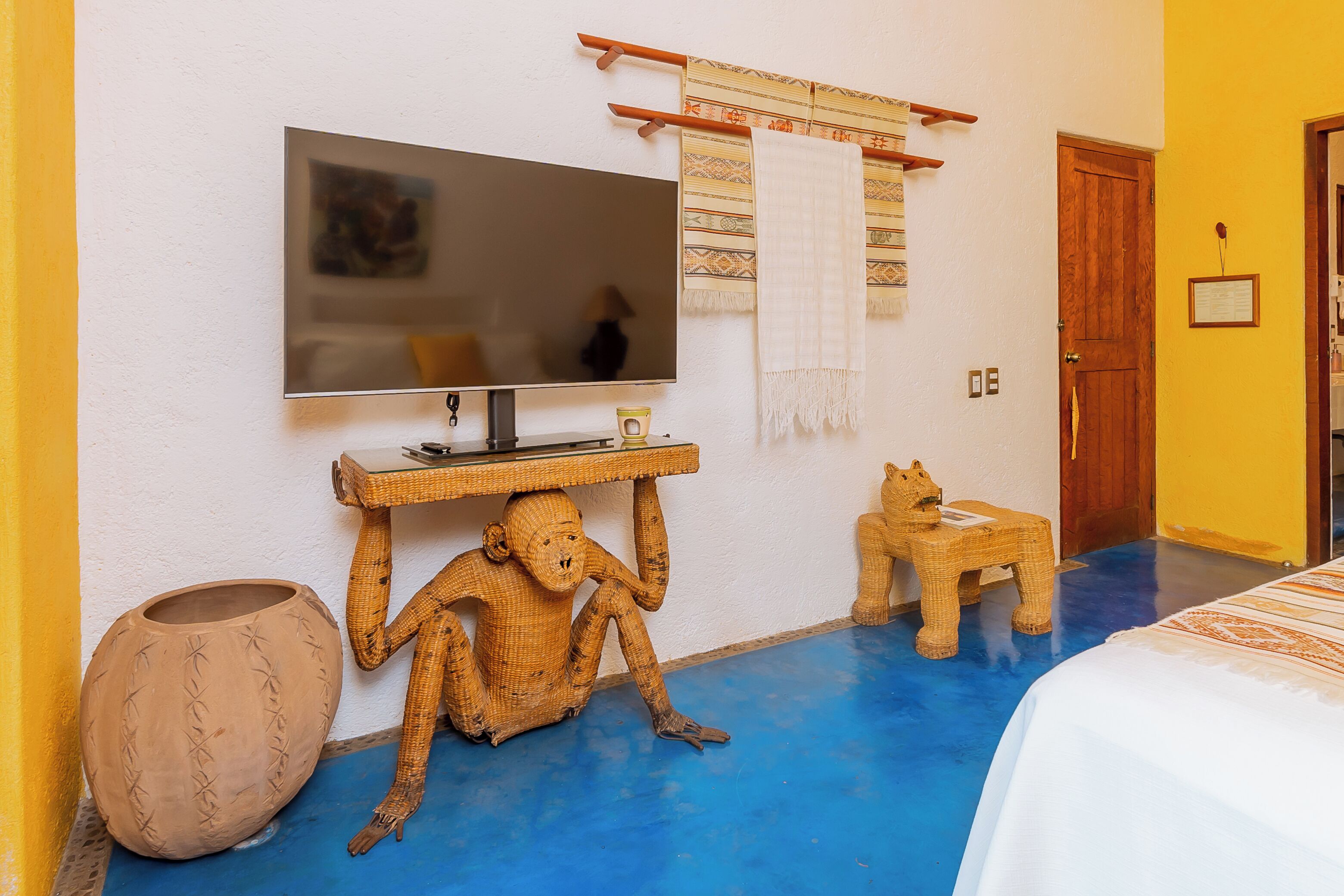 A room with a television mounted on the wall, a small table with a decorative item, a woven chair, a woven stool, and a large woven pot.