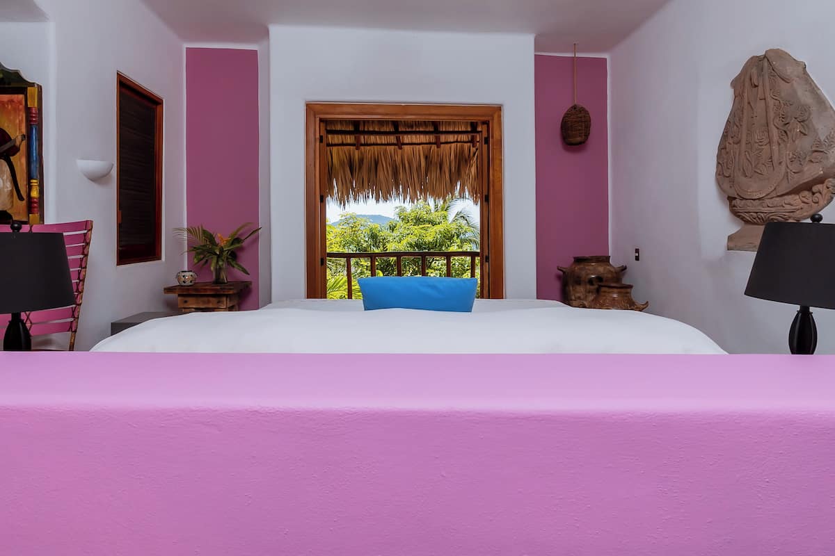 A bedroom with a large bed, a window with a view of greenery, a potted plant, and a decorative wall piece.