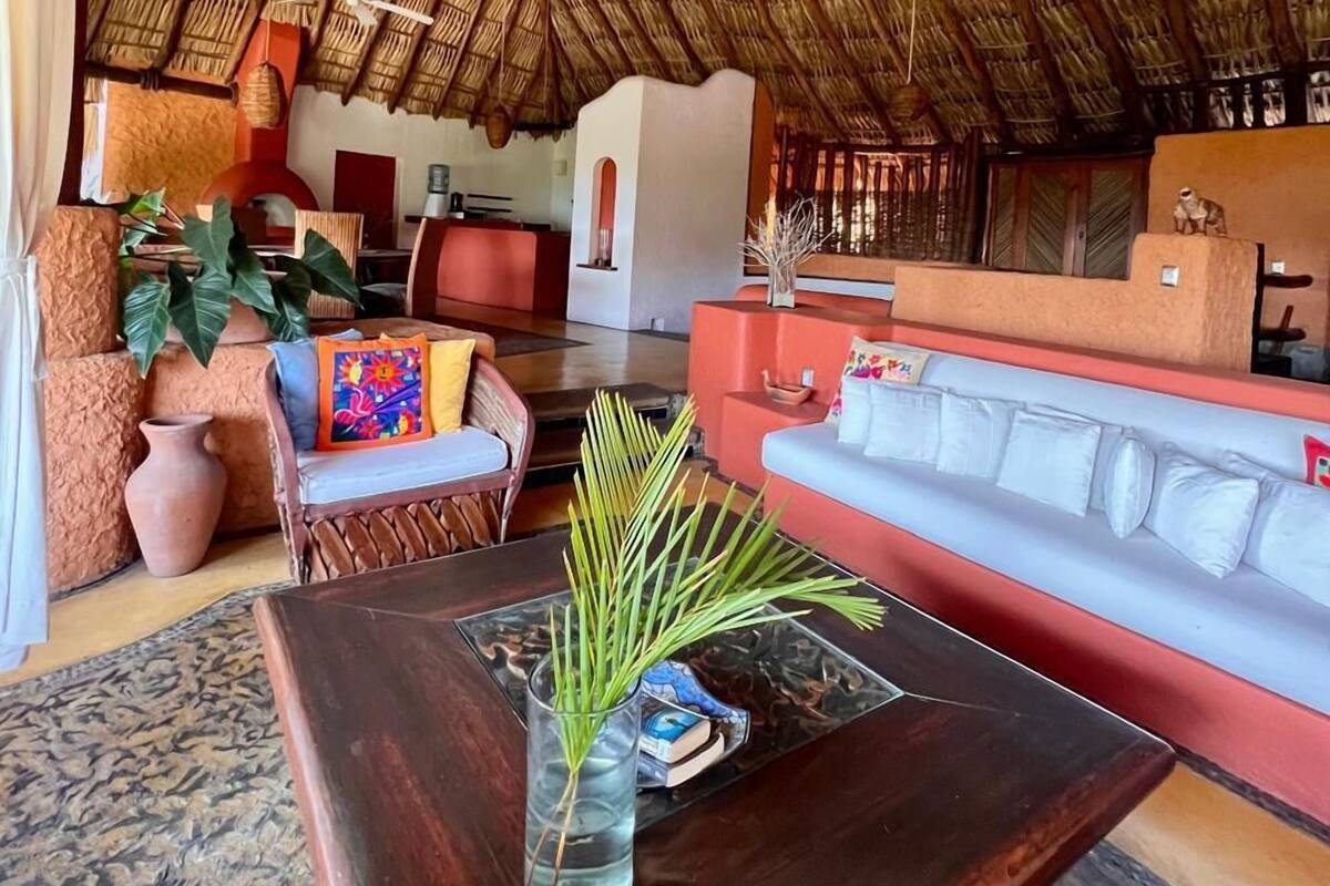 A spacious living area with a thatched roof, a wooden coffee table with a vase, a sofa, and a red couch.