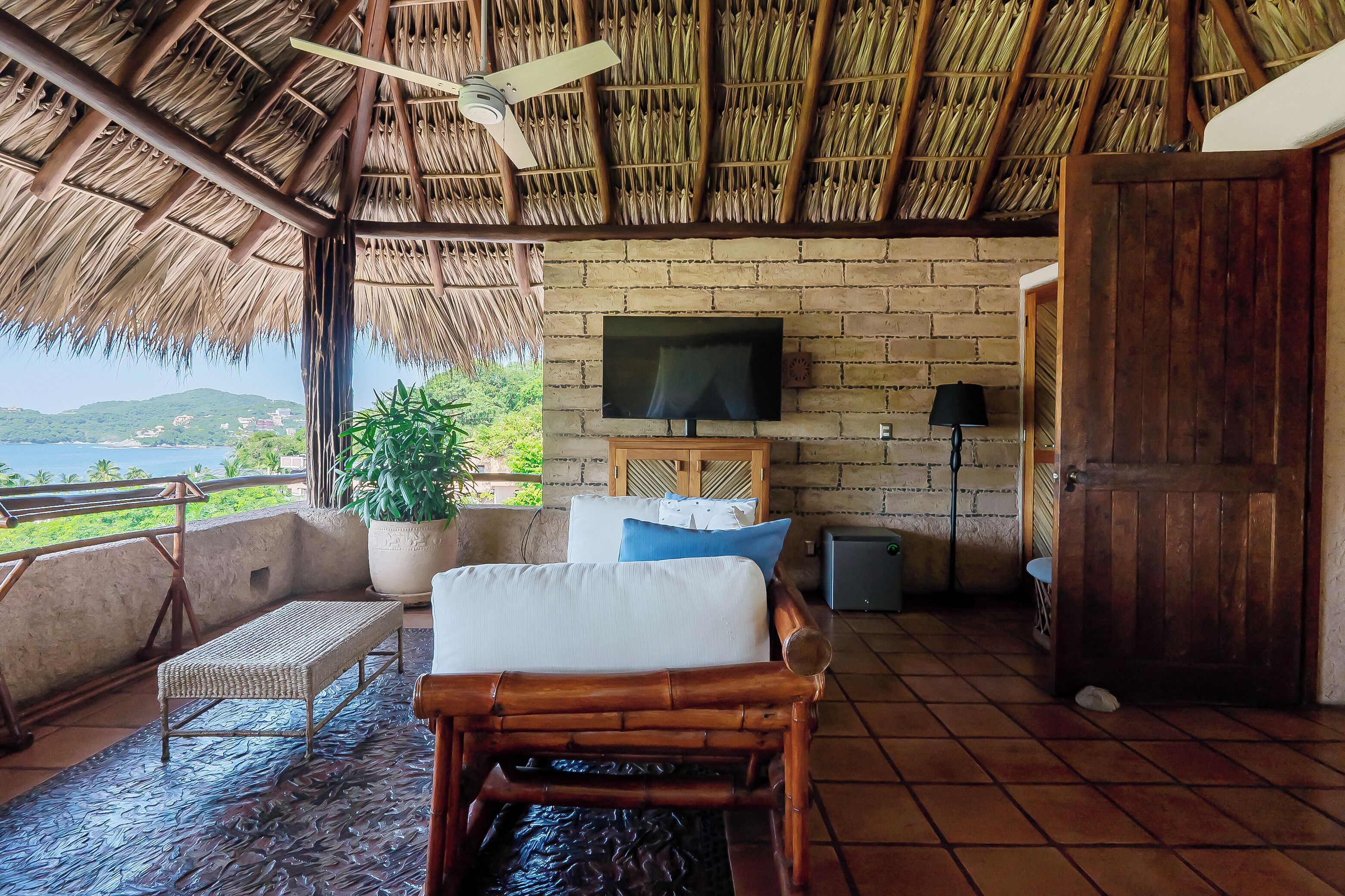 A room with a thatched roof, a TV mounted on a brick wall, a bench, a wooden chair, and a view of the sea.