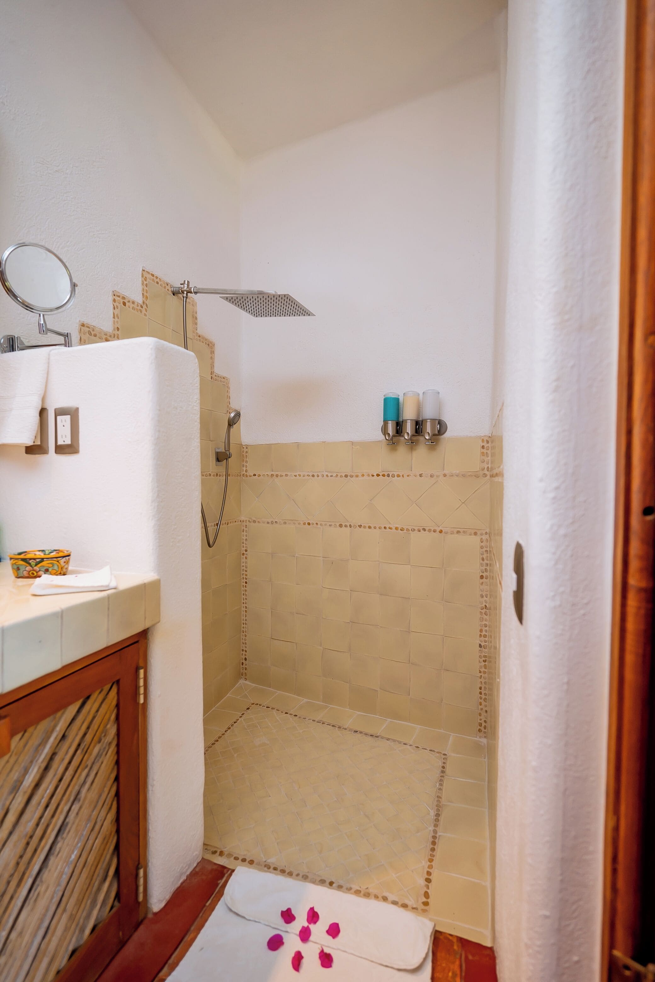 Un baño con ducha, lavamanos y espejo. Hay artículos de aseo personal en la pared y una toalla en el suelo.