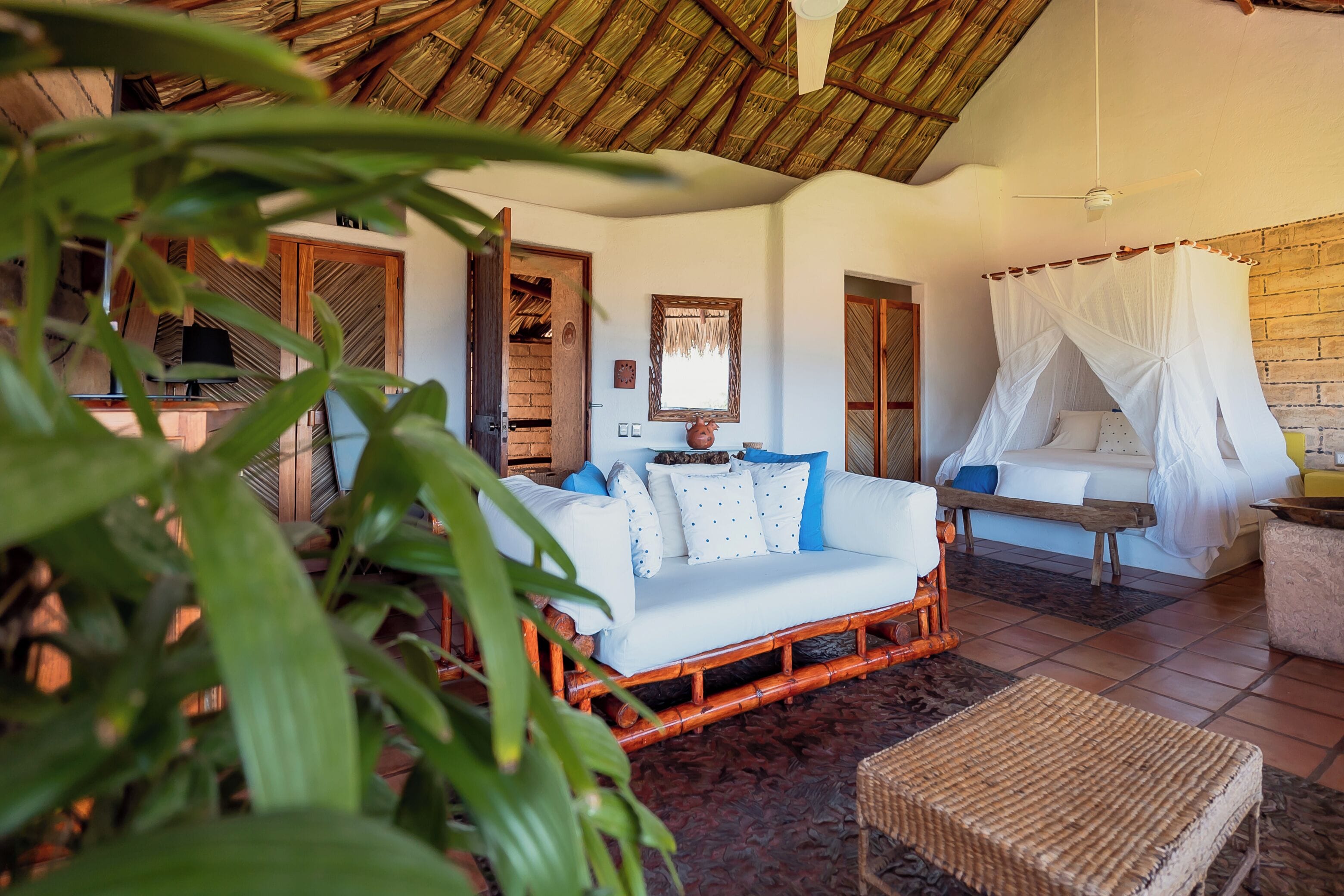 A room with a thatched roof, a white sofa with patterned cushions, a bed with a canopy, and a wicker chair.