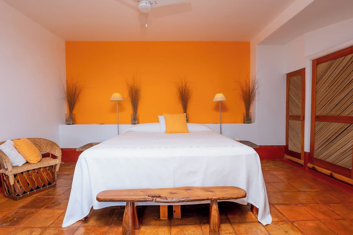 A bedroom with a large bed, a wooden bench, a wicker sofa, and two tall vases with branches.