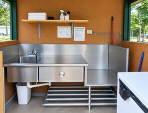 A compact kitchenette with a stainless steel sink, a trash bin, and a small shelf with cleaning supplies.