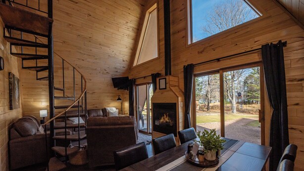 Een gezellig cabin interieur met een houten trap, een open haard, een eethoek en grote ramen met uitzicht op de natuur.