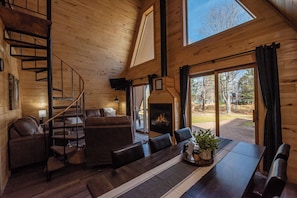 Interior acogedor de una cabaña con escalera de madera, chimenea, comedor y ventanas amplias con vistas al exterior.