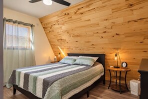 A bedroom with a sloped ceiling, wooden panel walls, a bed with striped bedding, two bedside lamps, and a small table with a clock.