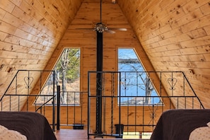 A wooden interior with a sloped ceiling, a central ceiling fan, and two windows offering views of trees and a body of water.