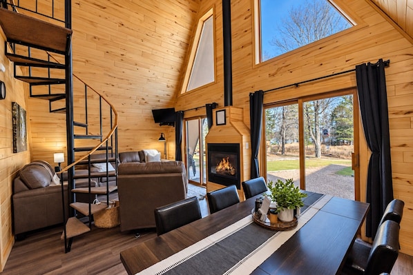 A cozy cabin interior with a fireplace, wooden walls, a sofa, a dining table, and a staircase.