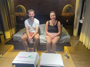 Two people sitting on a bed in a room with a YesHue logo, a bottle of water, and a small table. - YesHue Eco Waterfall Retreat (Hue)