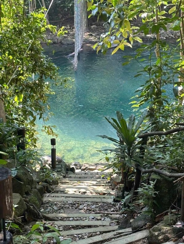 A stone pathway leading to a clear water body surrounded by lush greenery. - YesHue Eco Waterfall Retreat (Hue)