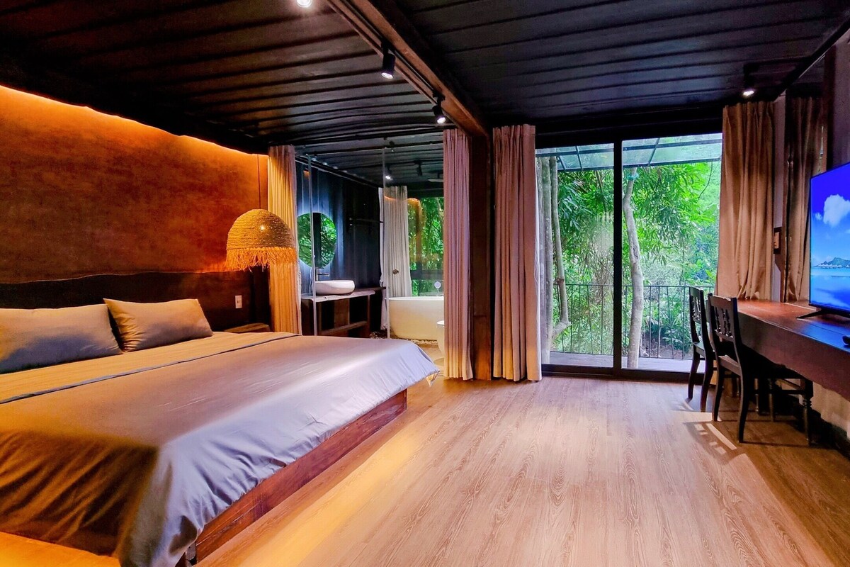 A modern bedroom with a large bed, a TV, and a balcony with a view of greenery.