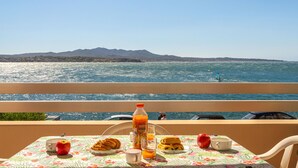 A table with breakfast items and a bottle of orange juice on a balcony overlooking the sea.