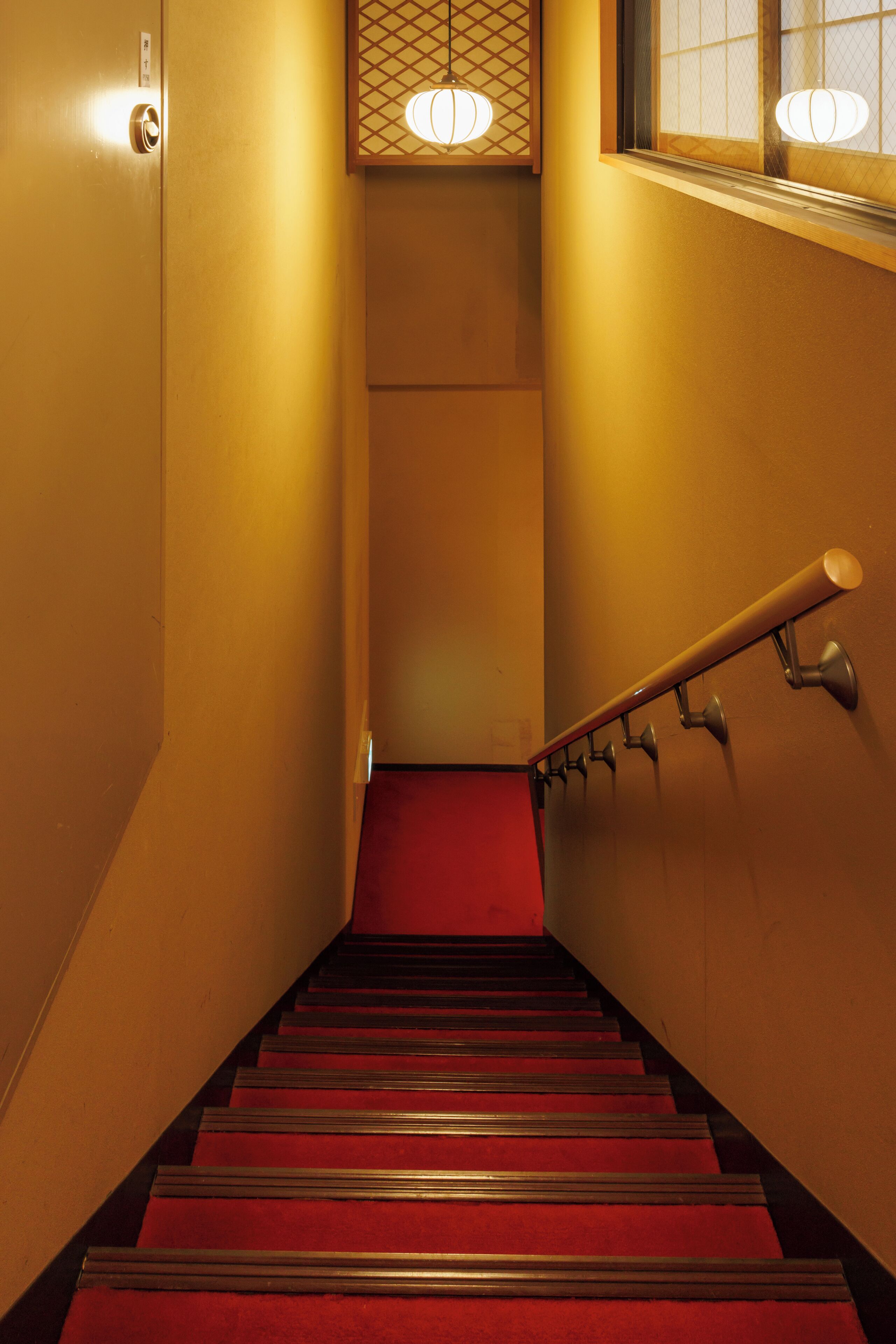 A staircase with a red carpet, a handrail, and a wall-mounted light fixture.