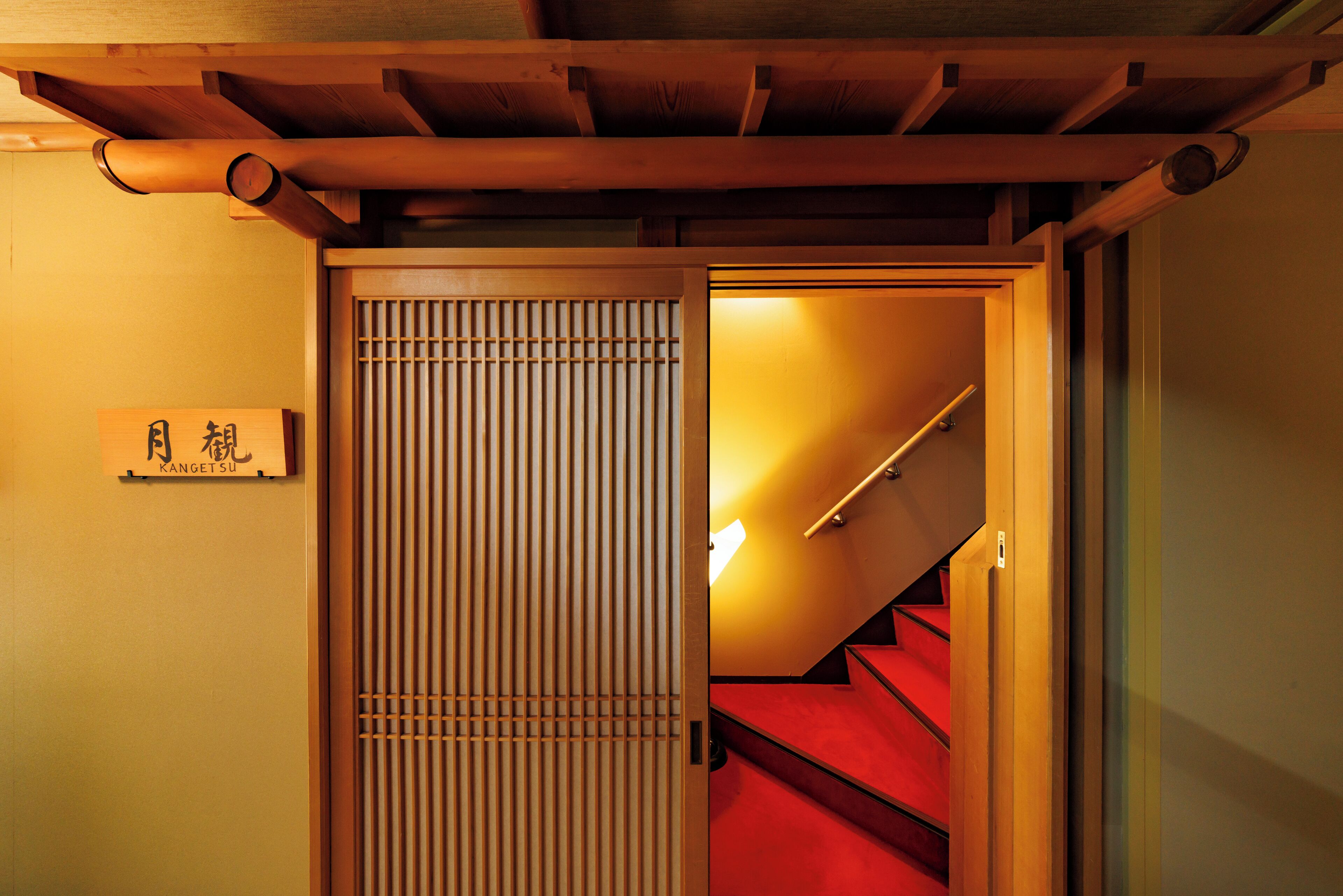 A traditional Japanese interior with a wooden sliding door, a staircase with red steps, and a sign with Japanese characters.