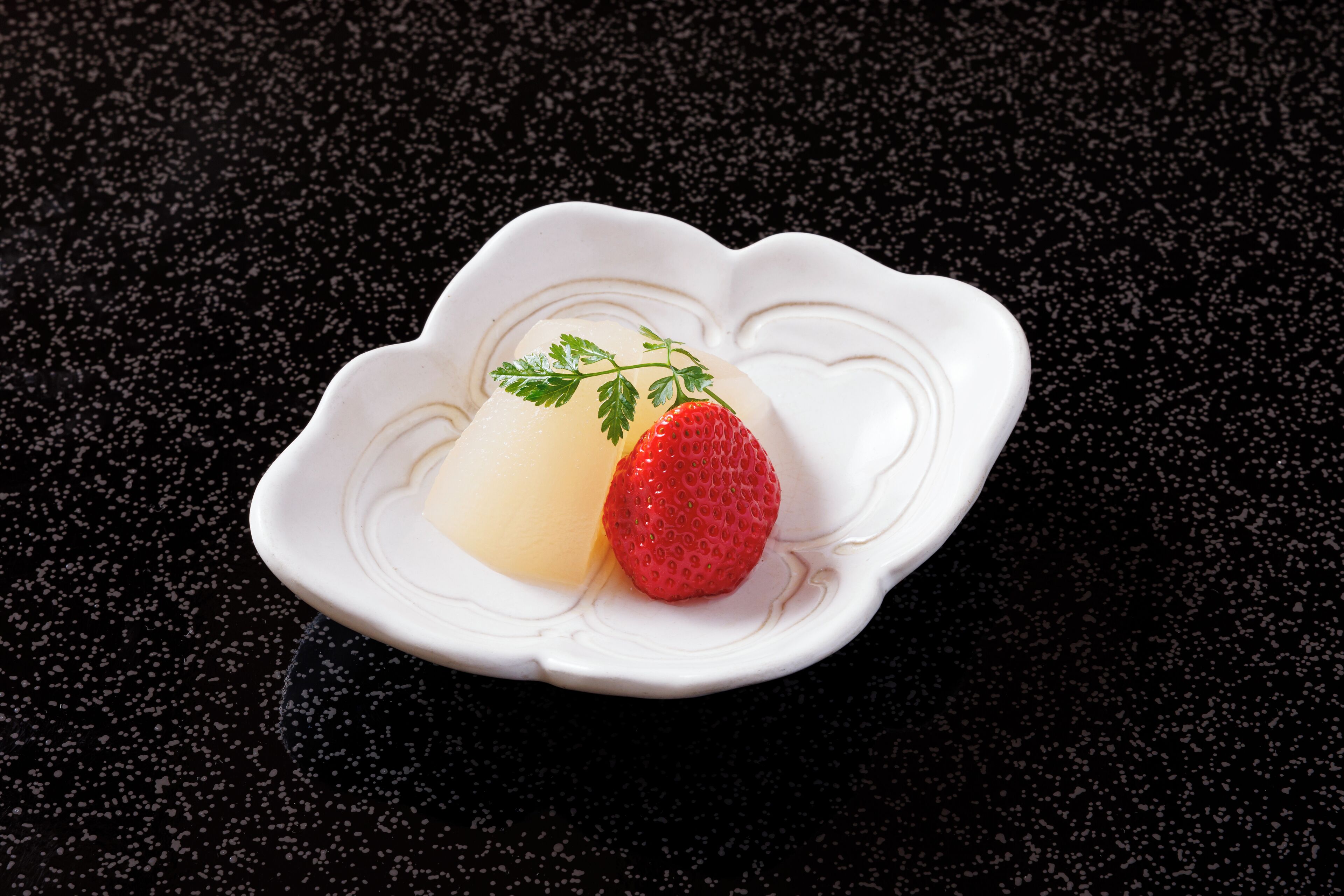 A white dessert plate with a single strawberry and a pale dessert on a dark surface.