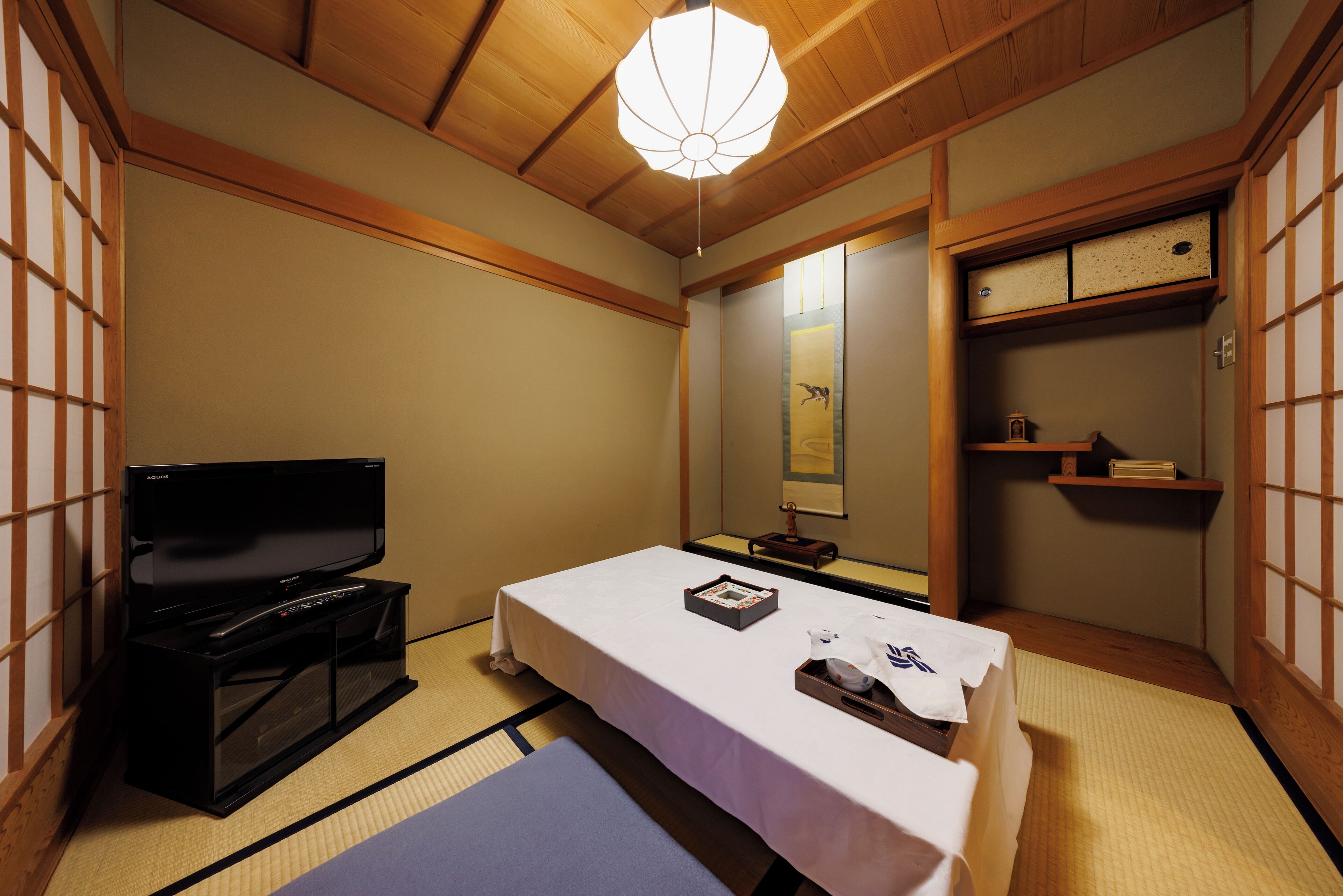 Een traditionele Japanse kamer met tatami vloeren, een televisie, een bed met een witte spreidek en een plank met een spiegel en kleine objecten.