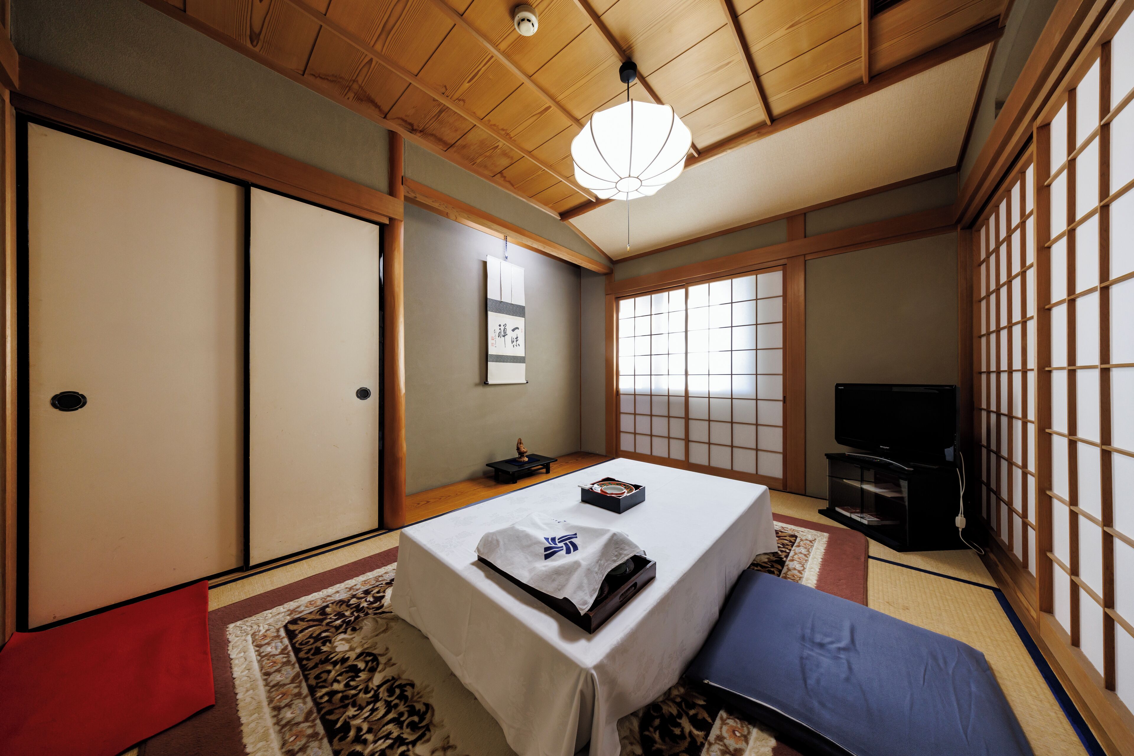 A traditional Japanese room with tatami flooring, a low table with a white tablecloth, a television, and sliding doors.