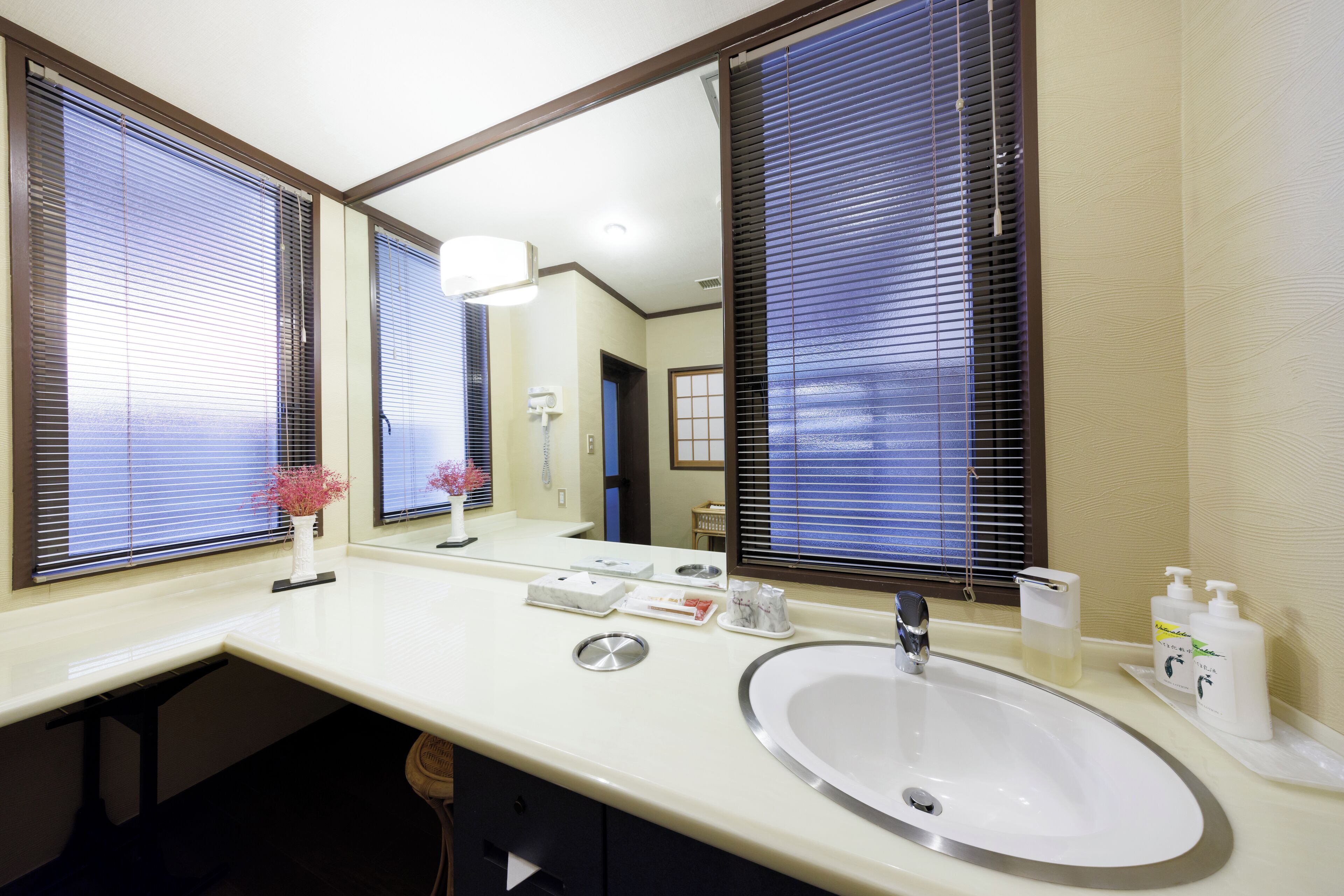 A hotel bathroom with a large mirror, a sink, and a window with blinds.