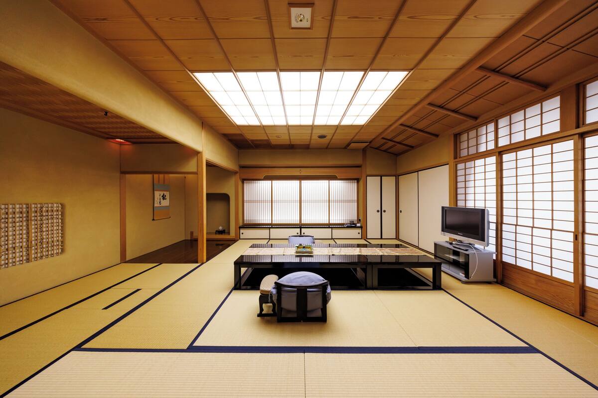 A traditional Japanese room with tatami flooring, a tatami mat table, a low table, a television, and sliding doors.