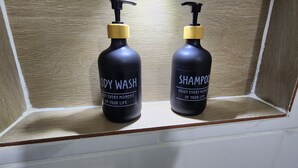 Kaksi mustaa pulloa, joissa on keltaiset pumppupullot, ja joissa lukee etiketissä ”KAPPALPESU” ja ”SHAMPOO”, on asetettu puuhyllylle.