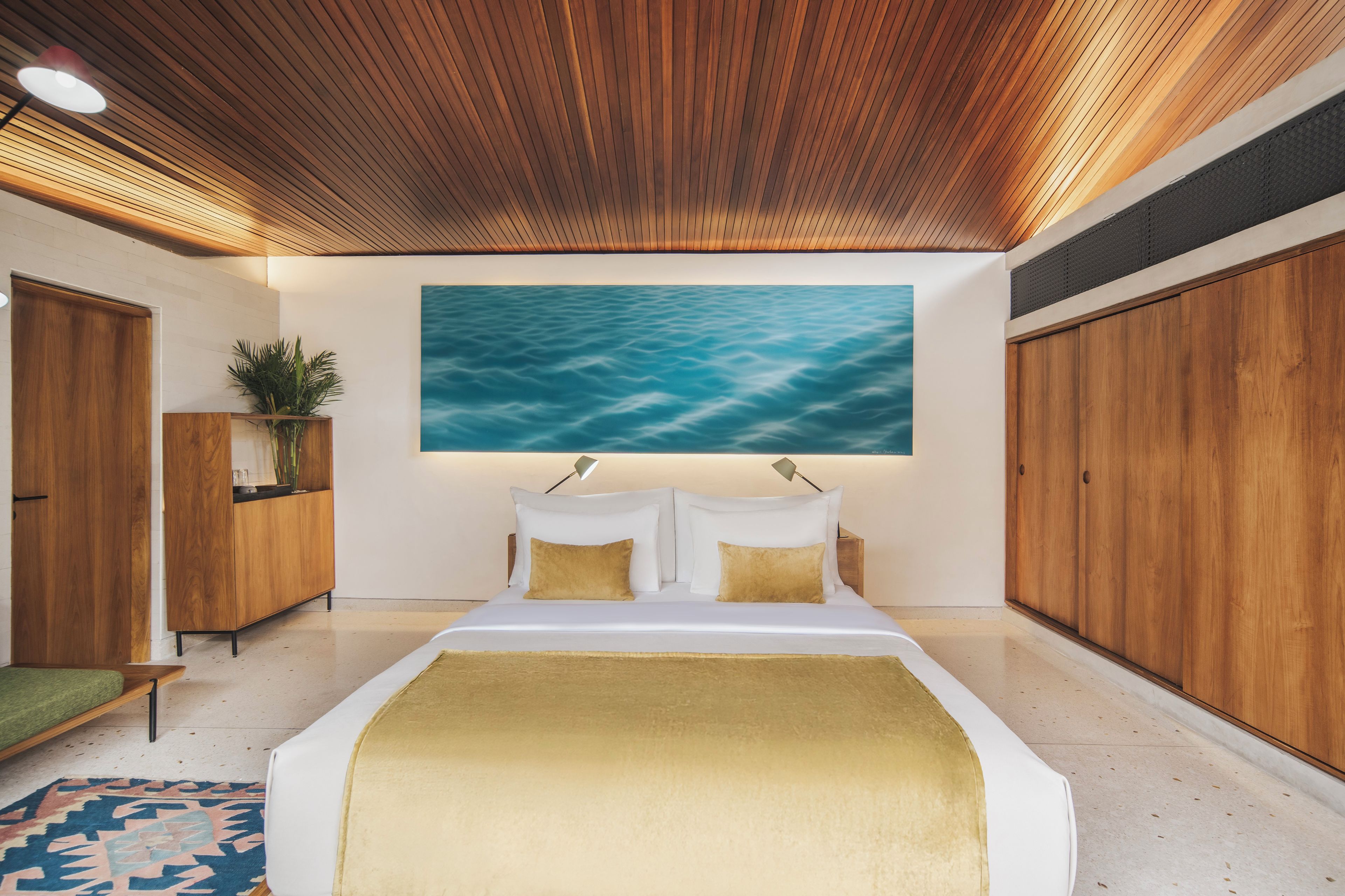 A modern bedroom with a wooden ceiling, a large bed with white bedding, a painting of water on the wall, and a green rug.