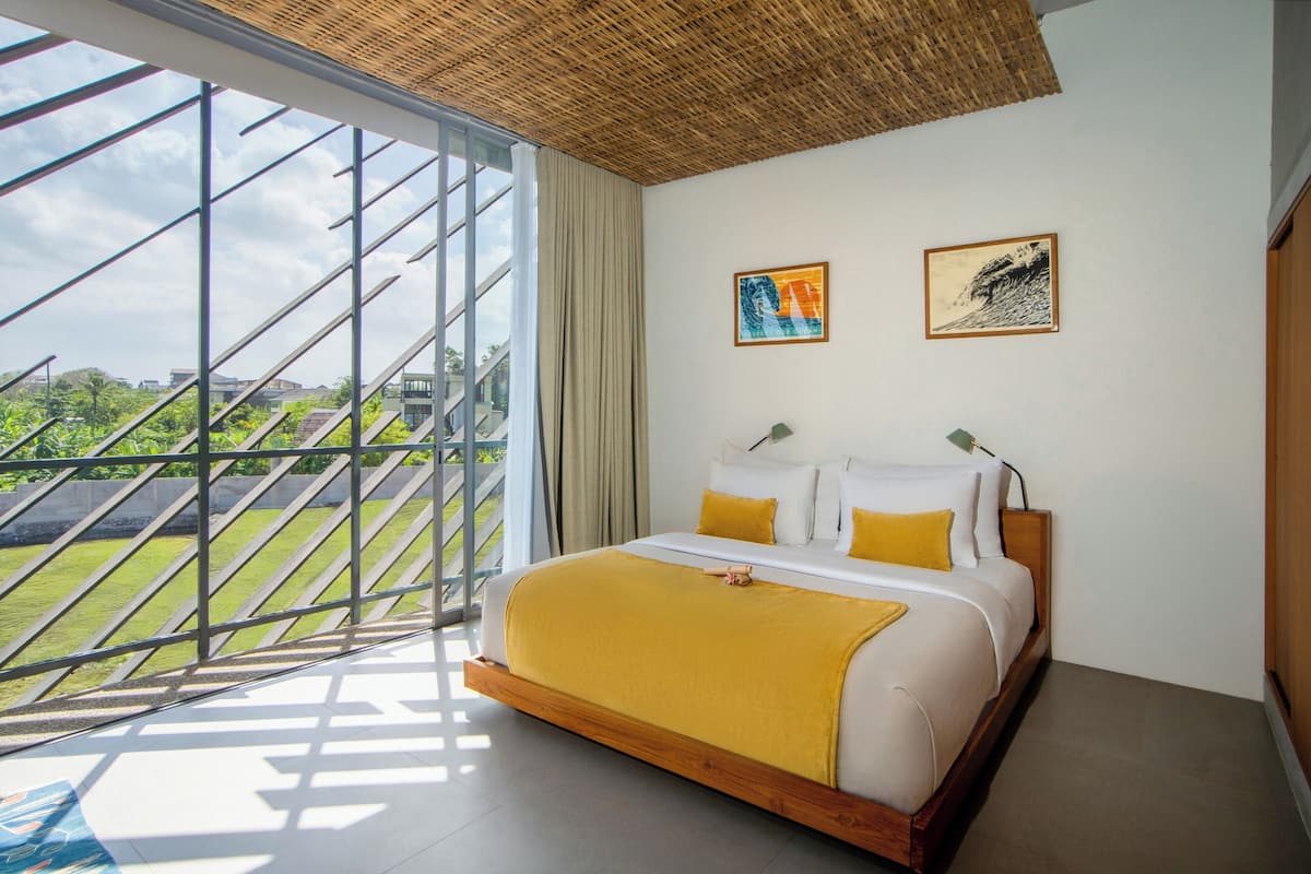 Een slaapkamer met een groot raam, een bed met wit en geel beddengoed, en twee kunstwerken in een lijst aan de muur.