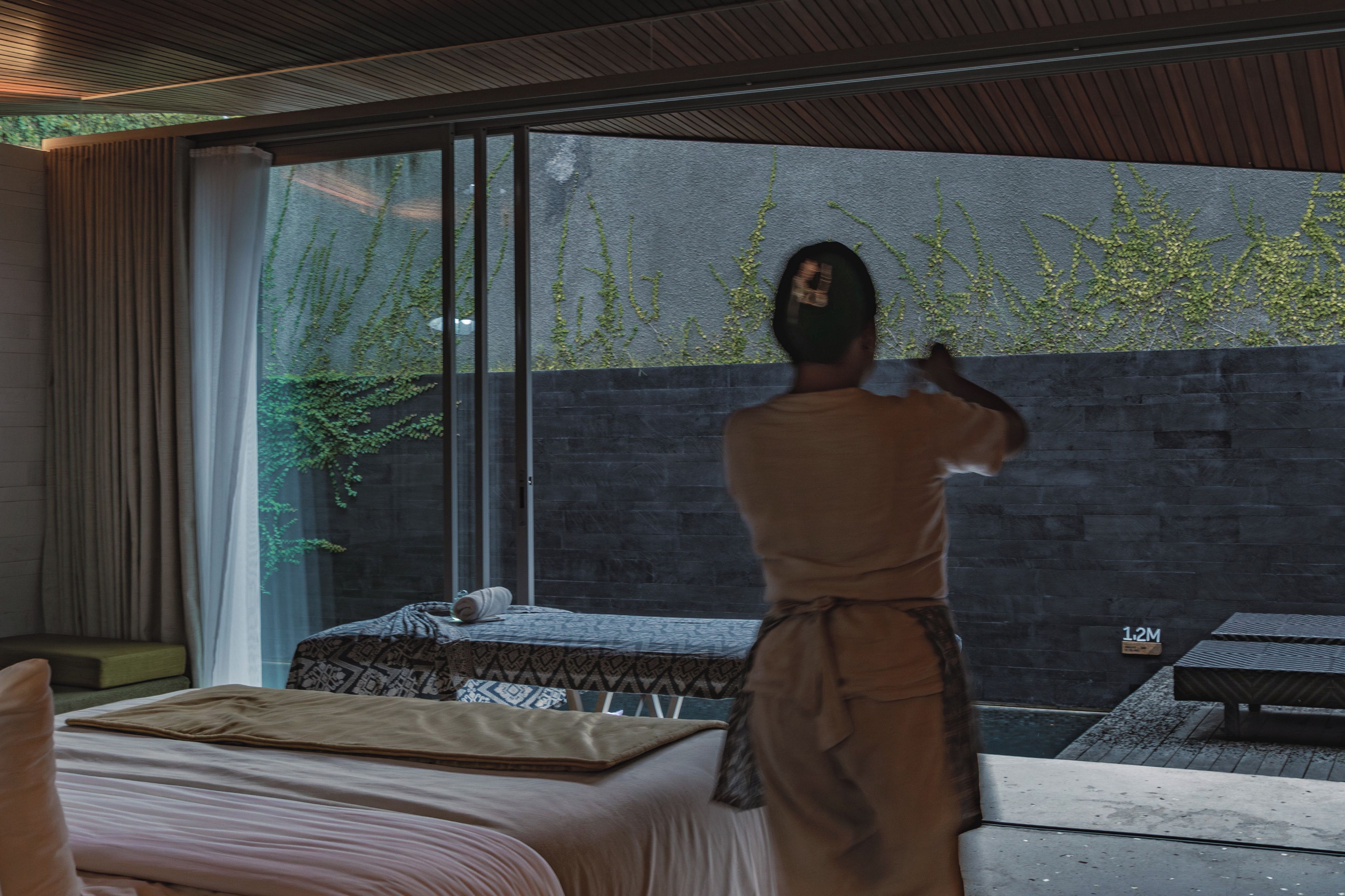 A person in a robe standing by a window with a view of a stone wall and greenery outside.