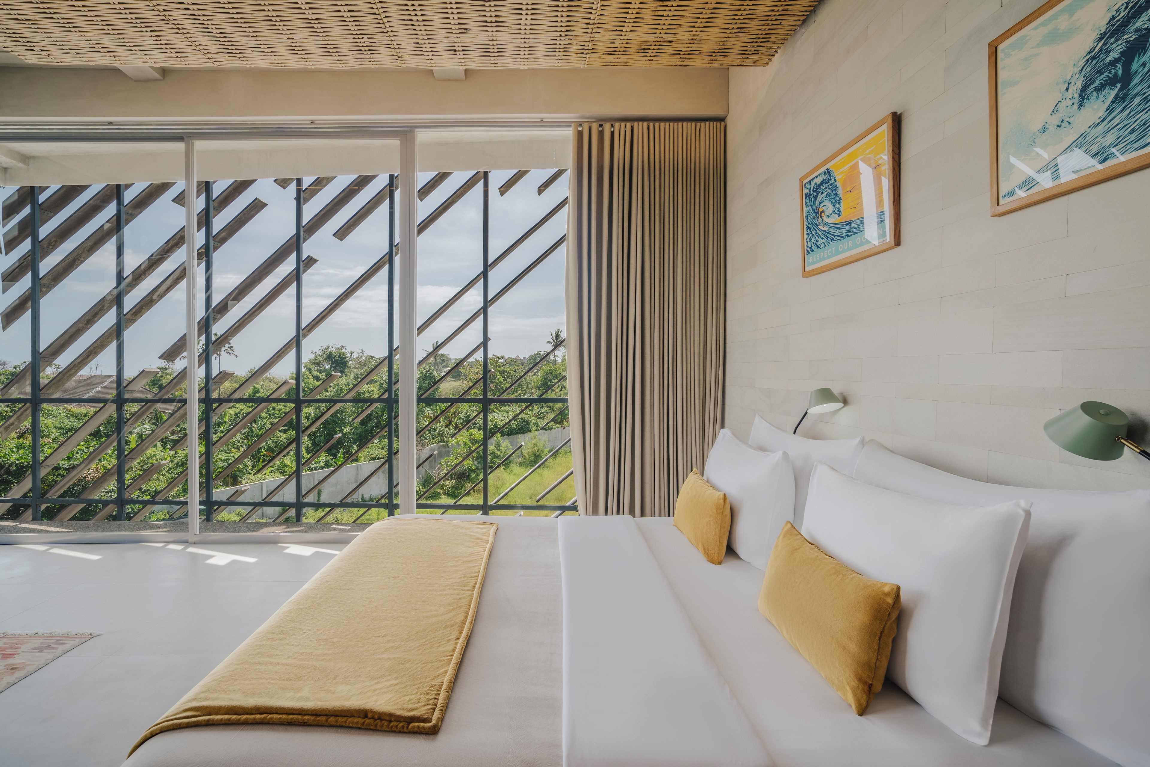 Quarto de hotel com uma janela grande, cama com roupa de cama branca e um cobertor bege, e abajur verde na parede.