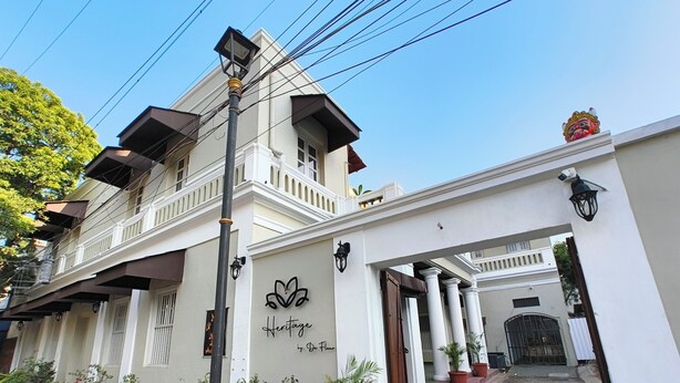 Un edificio con fachada blanca, un letrero que dice "Heritage by De Fleur" y una serie de macetas con plantas a lo largo de la entrada.