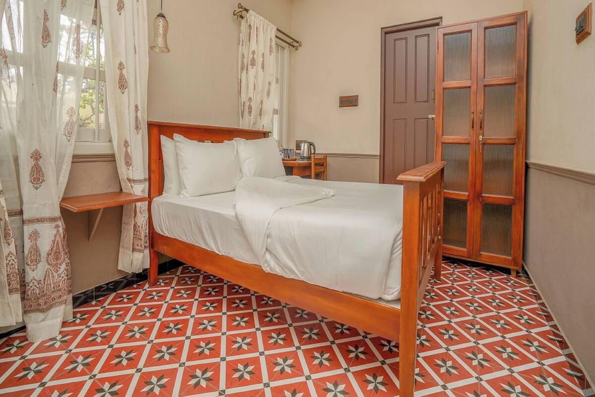 A bedroom with a wooden bed, a wardrobe, a small table with a kettle, and patterned floor tiles.