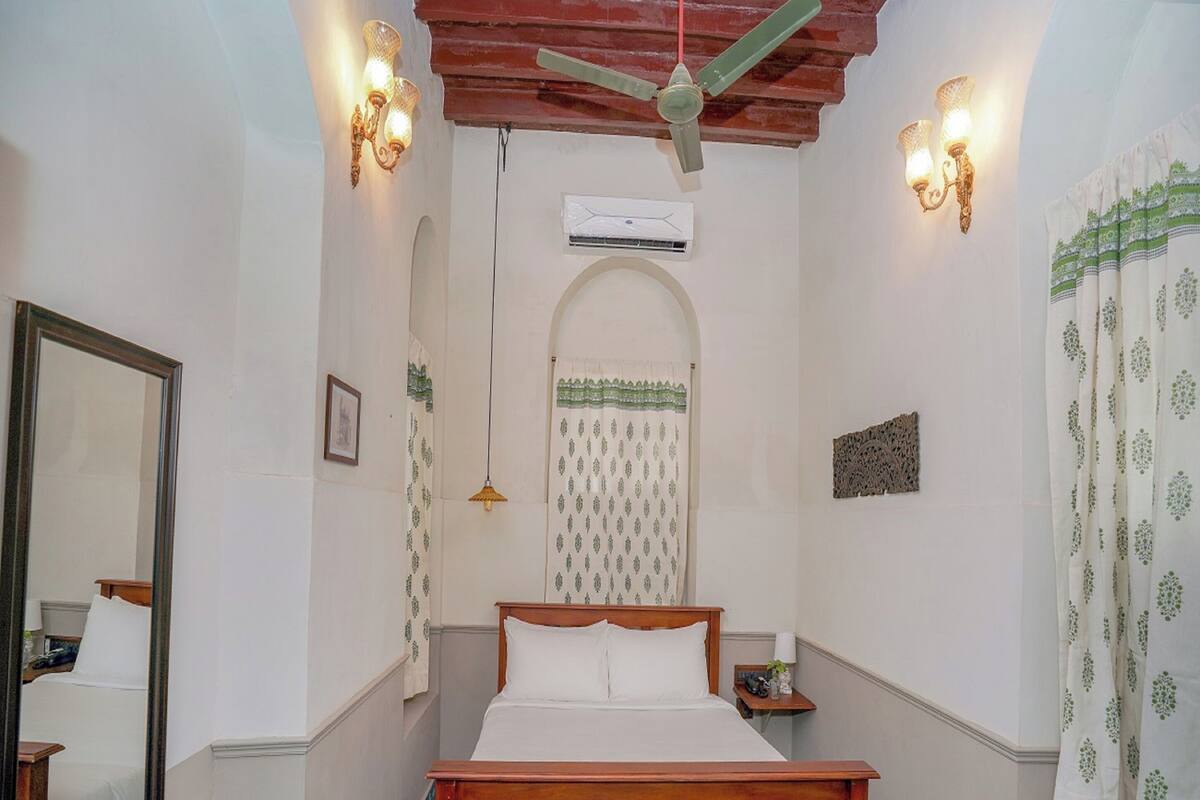 A bedroom with a wooden bed, a ceiling fan, wall-mounted lights, a mirror, and patterned curtains.