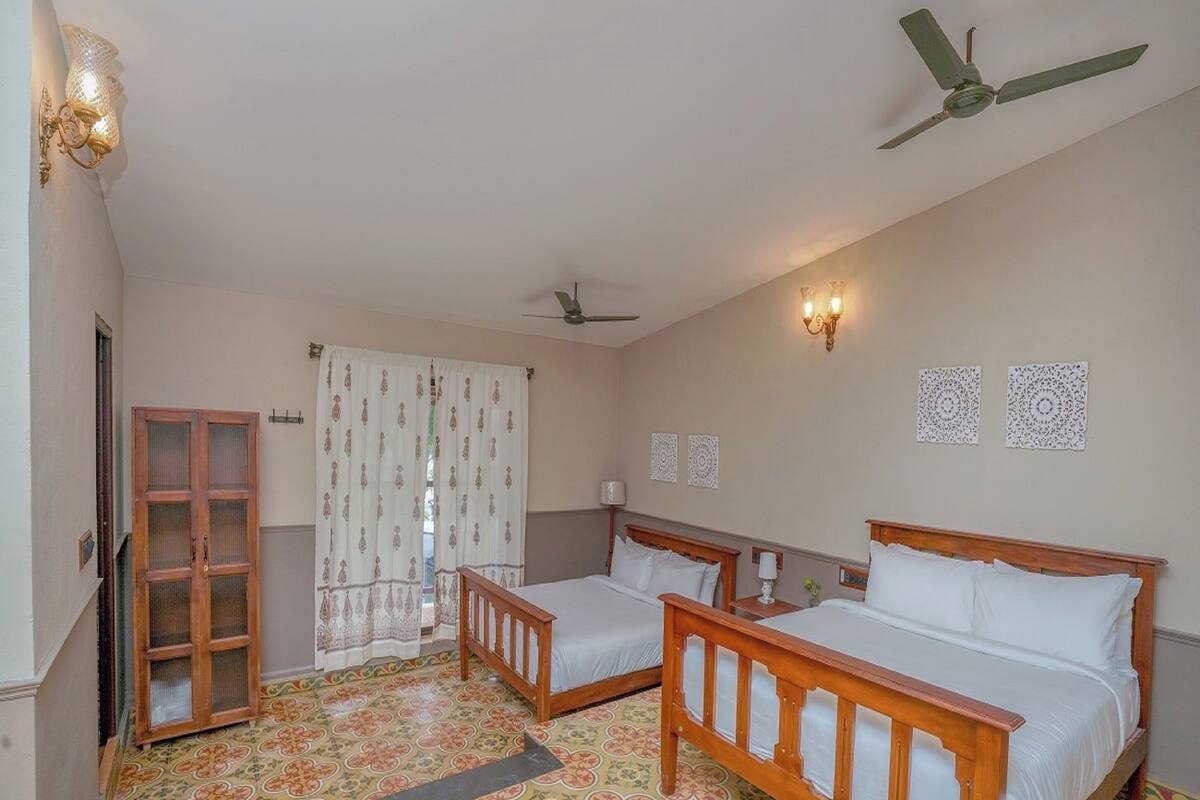 A room with two wooden beds, a ceiling fan, a window with patterned curtains, and a door with a glass panel.