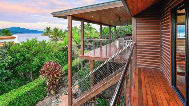 A wooden deck with a glass balustrade overlooking a lush garden and a body of water.