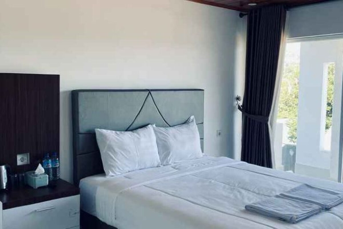 A hotel room with a bed, a nightstand, a window with curtains, and a view of greenery outside.