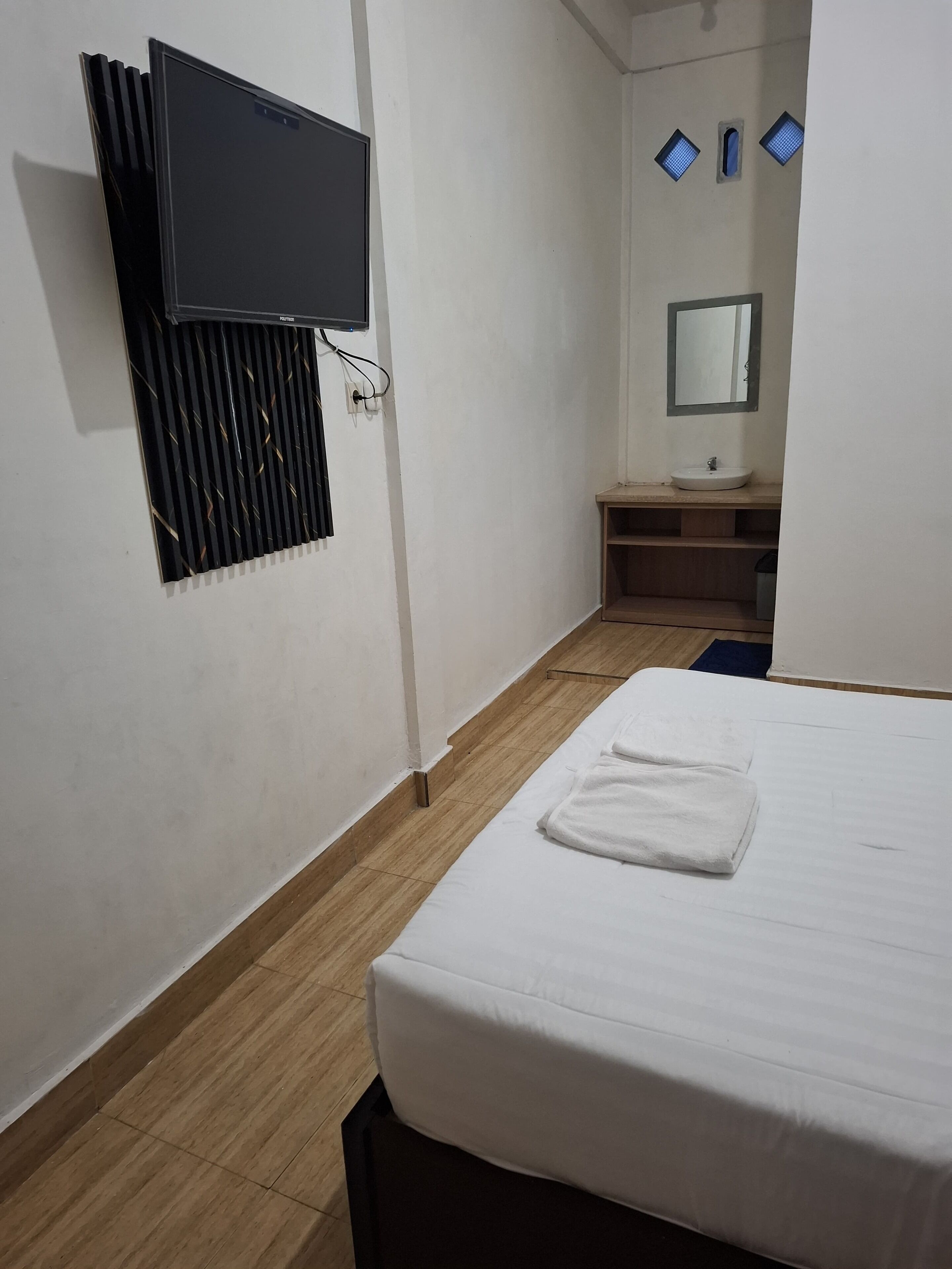 A hotel room with a bed, a TV mounted on the wall, a small bathroom with a sink and mirror, and a window with blinds.