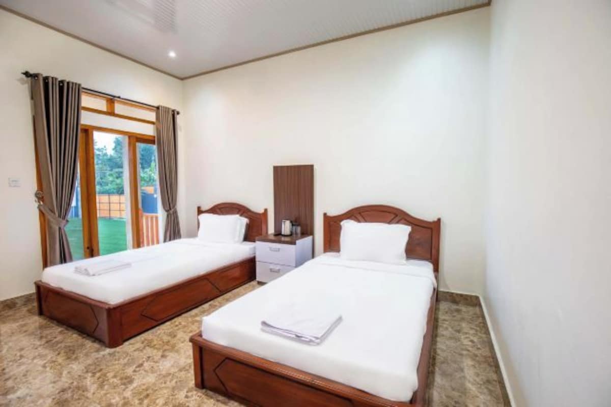 Habitación de hotel con dos camas de madera, una mesita de noche con lámpara y vistas a un espacio exterior verde a través de una ventana con cortinas.