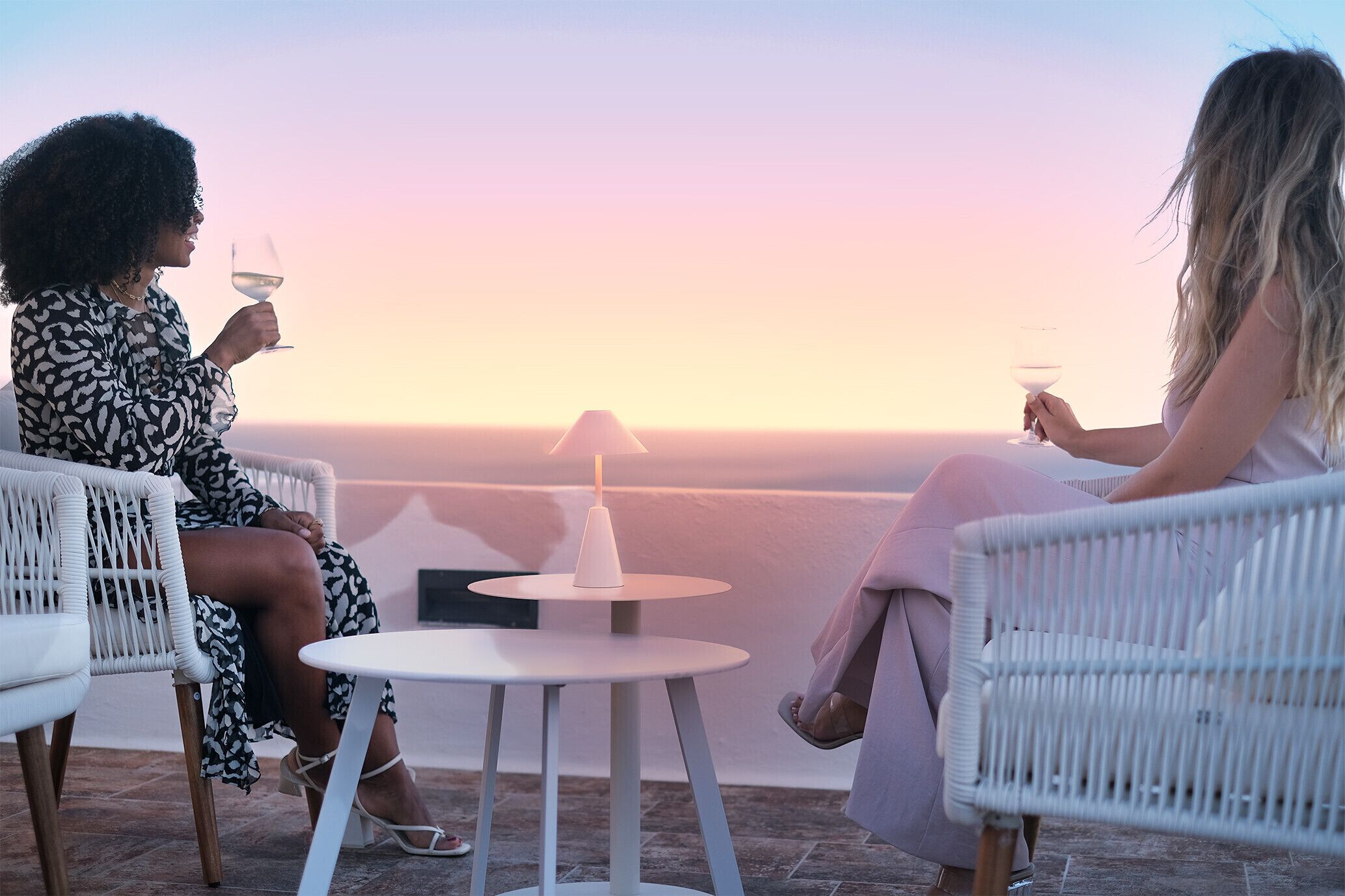 Two people sitting on white wicker chairs at a small round table with a white table lamp, overlooking a scenic view during sunset.