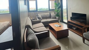 A modern living room with a sofa set, a coffee table, a TV stand, and a large window with a view. - Executive 1BR One Manchester Place (Lapu-Lapu)