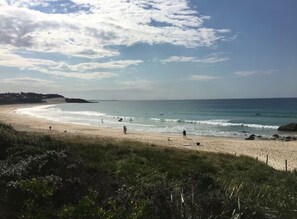 A beach with a curved shoreline, people walking along the shore, and a clear sky with scattered clouds. - Beachfront house on beautiful One Mile Beach (Forster)