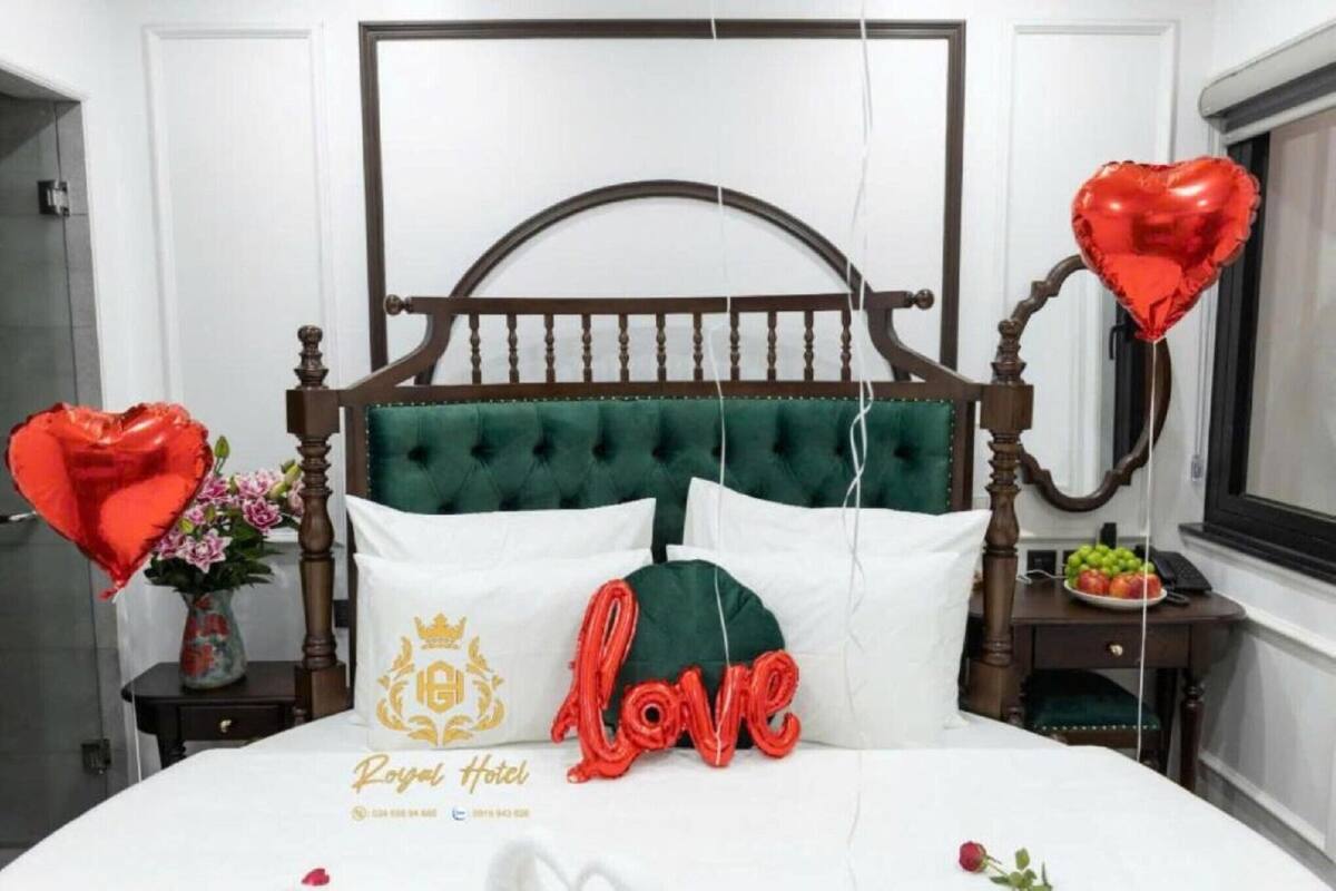 Una habitación de hotel con cama, mesita de noche, un jarrón con flores y globos con forma de corazón.