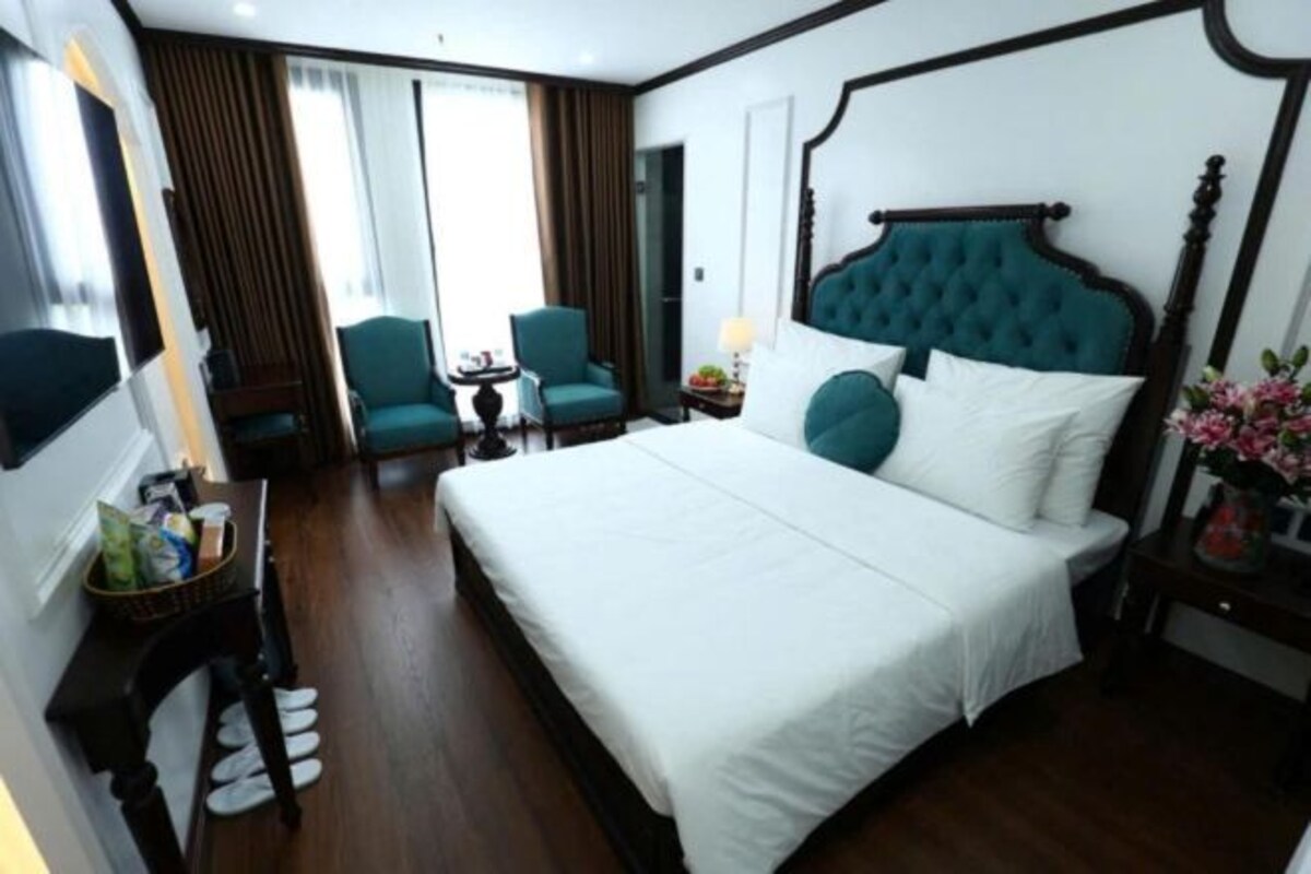 A hotel room with a large bed, a desk with a chair, a small table with a lamp, and a TV on the wall.