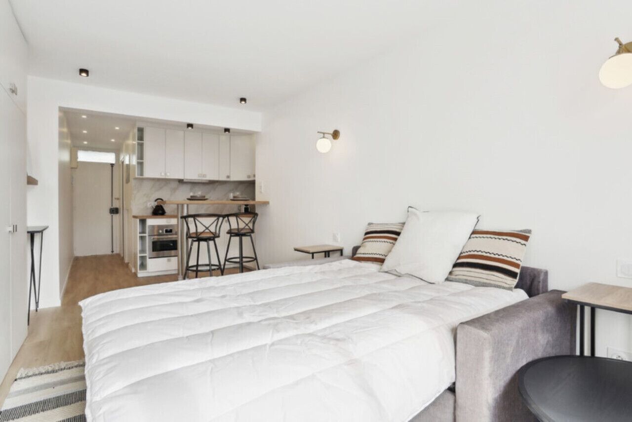 A modern, compact bedroom with a large bed, a small desk, and a kitchenette in the background.