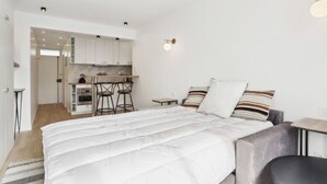 A modern, compact bedroom with a large bed, a small desk, and a kitchenette in the background. - Bail mobilité - grand studio - Paris (Boulogne-Billancourt)