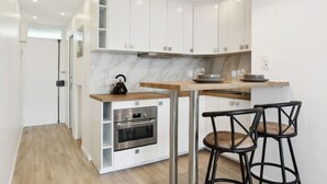 A modern kitchen with white cabinets, a marble countertop, and a built-in oven. There are two black bar stools and a small island with a kettle. - Bail mobilité - grand studio - Paris (Boulogne-Billancourt)