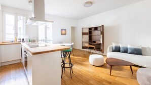 A modern kitchen with a central island, a dining area with a table and chairs, a sofa, and a bookshelf.