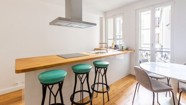 A modern kitchen with a wooden countertop, bar stools, a round dining table, and a view of the city through large windows.