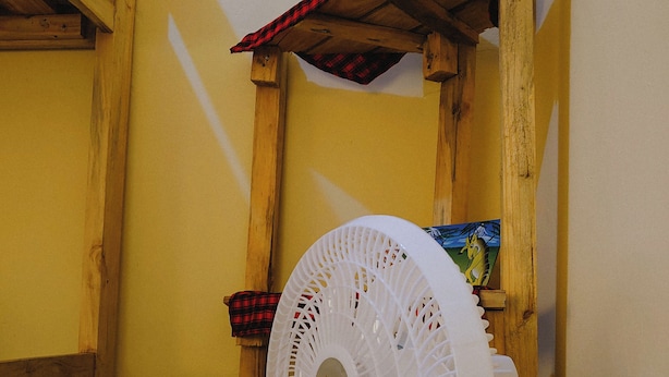A room with a wooden ceiling structure, a white standing fan, and a small shelf with a box. - Sero Safaris Hostel (Arusha)