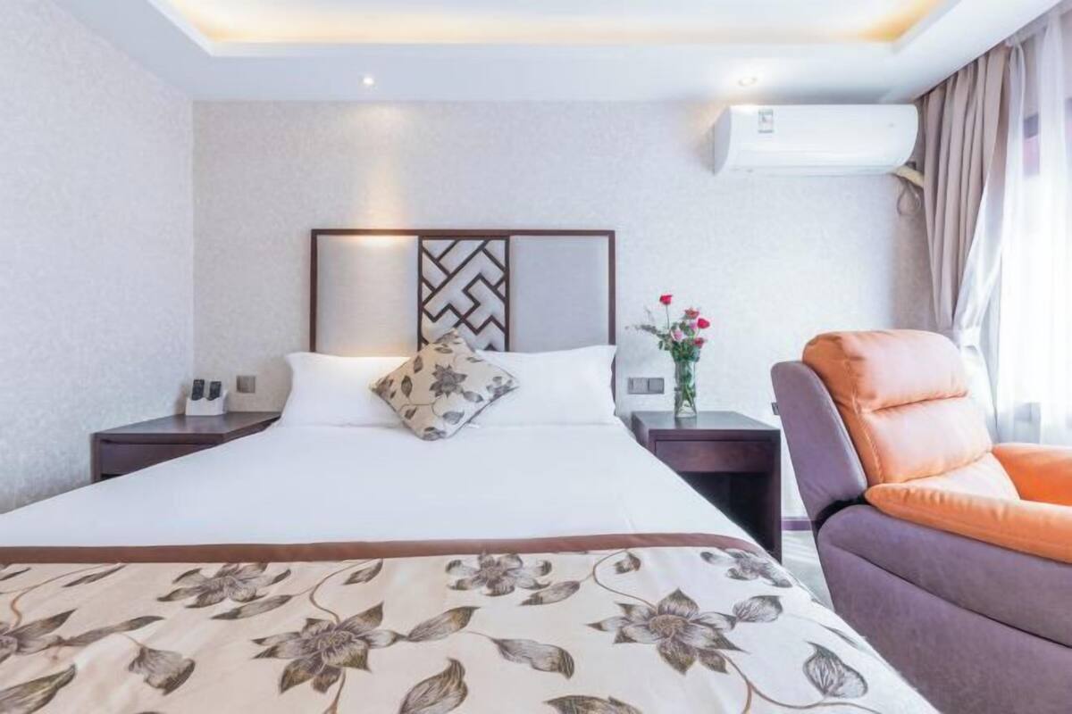 A hotel room with a bed, a nightstand, a chair, a decorative wall panel, and an air conditioning unit.